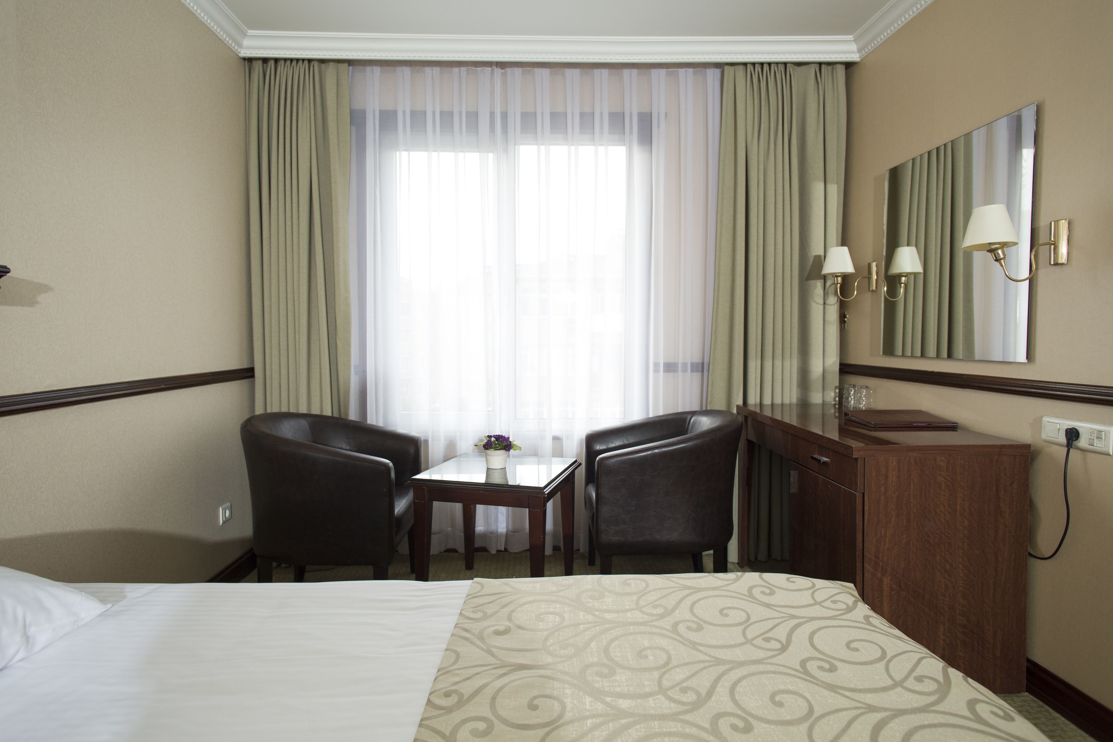A hotel room with a bed, two chairs, a small table with a vase, a mirror, and a window with curtains.