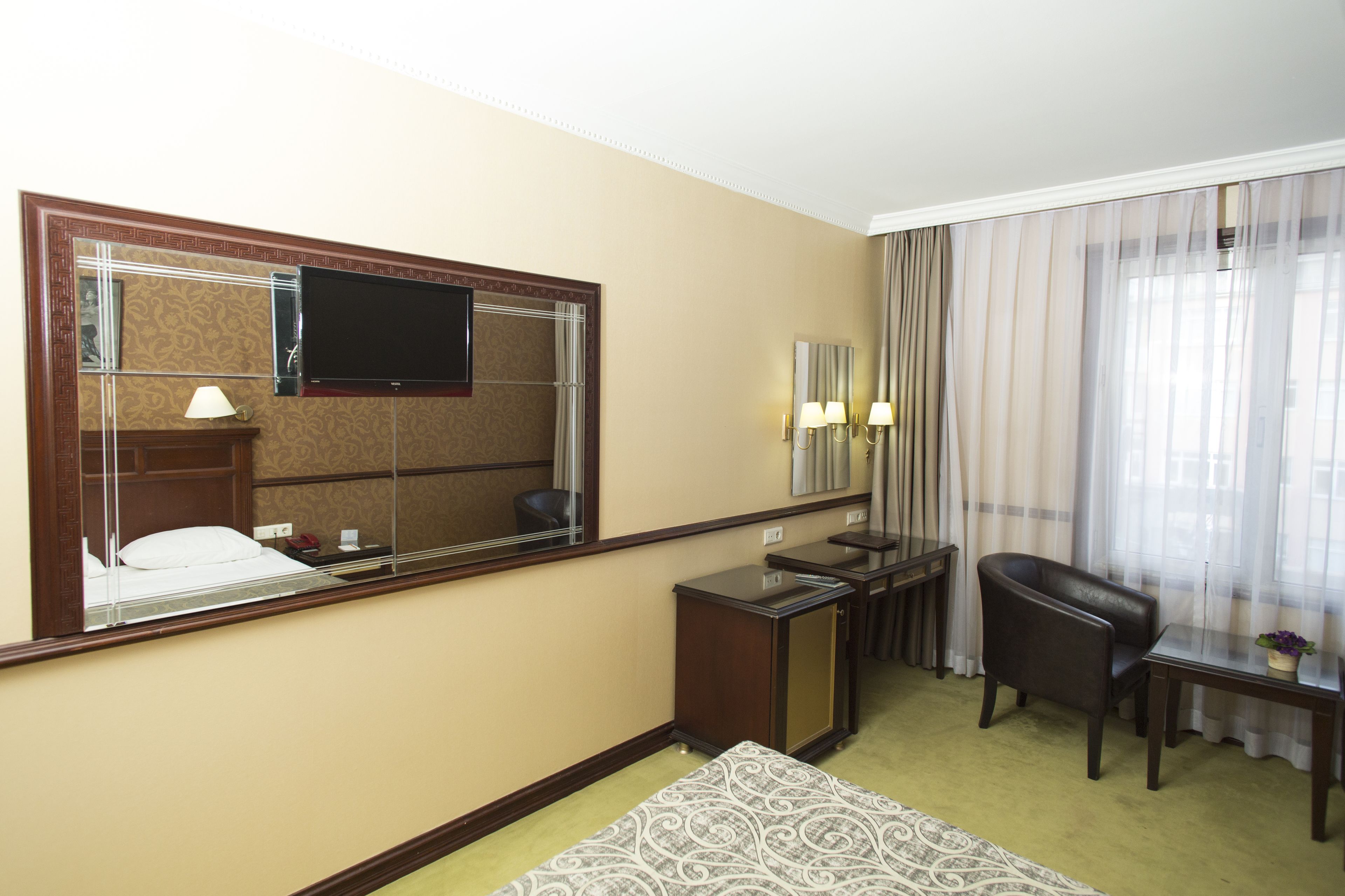 A hotel room with a large mirror, a flat-screen TV, a desk with two lamps, a leather chair, and a small table with a plant.