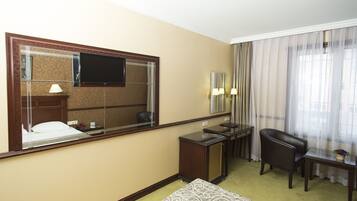 A hotel room with a large mirror, a flat-screen TV, a desk with two lamps, a leather chair, and a small table with a plant.