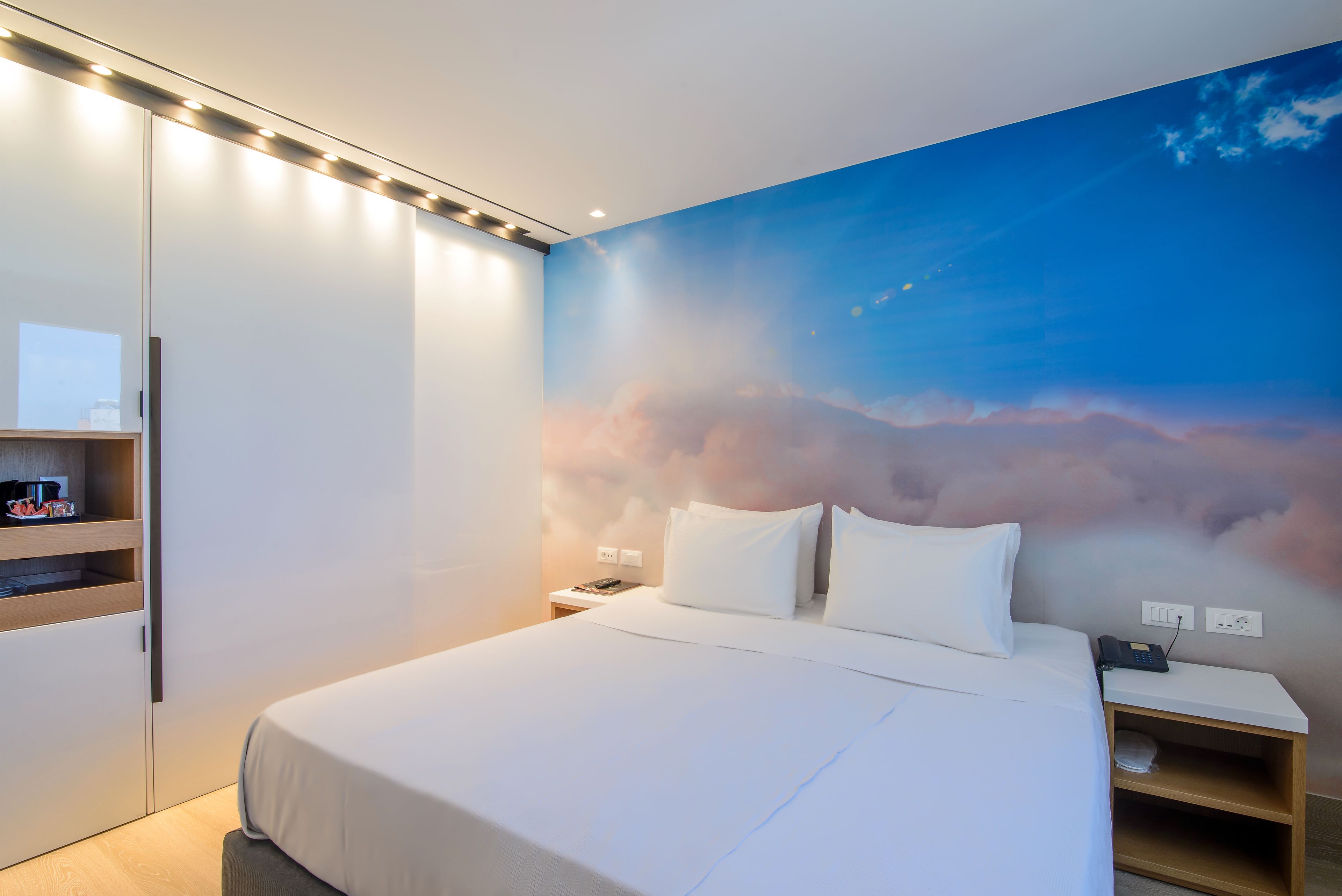 Una habitación de hotel moderna con una cama grande, una mesita de noche, un armario y un mural en la pared que muestra un cielo con nubes.