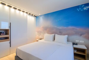 Una habitación de hotel moderna con una cama grande, una mesita de noche, un armario y un mural en la pared que muestra un cielo con nubes.