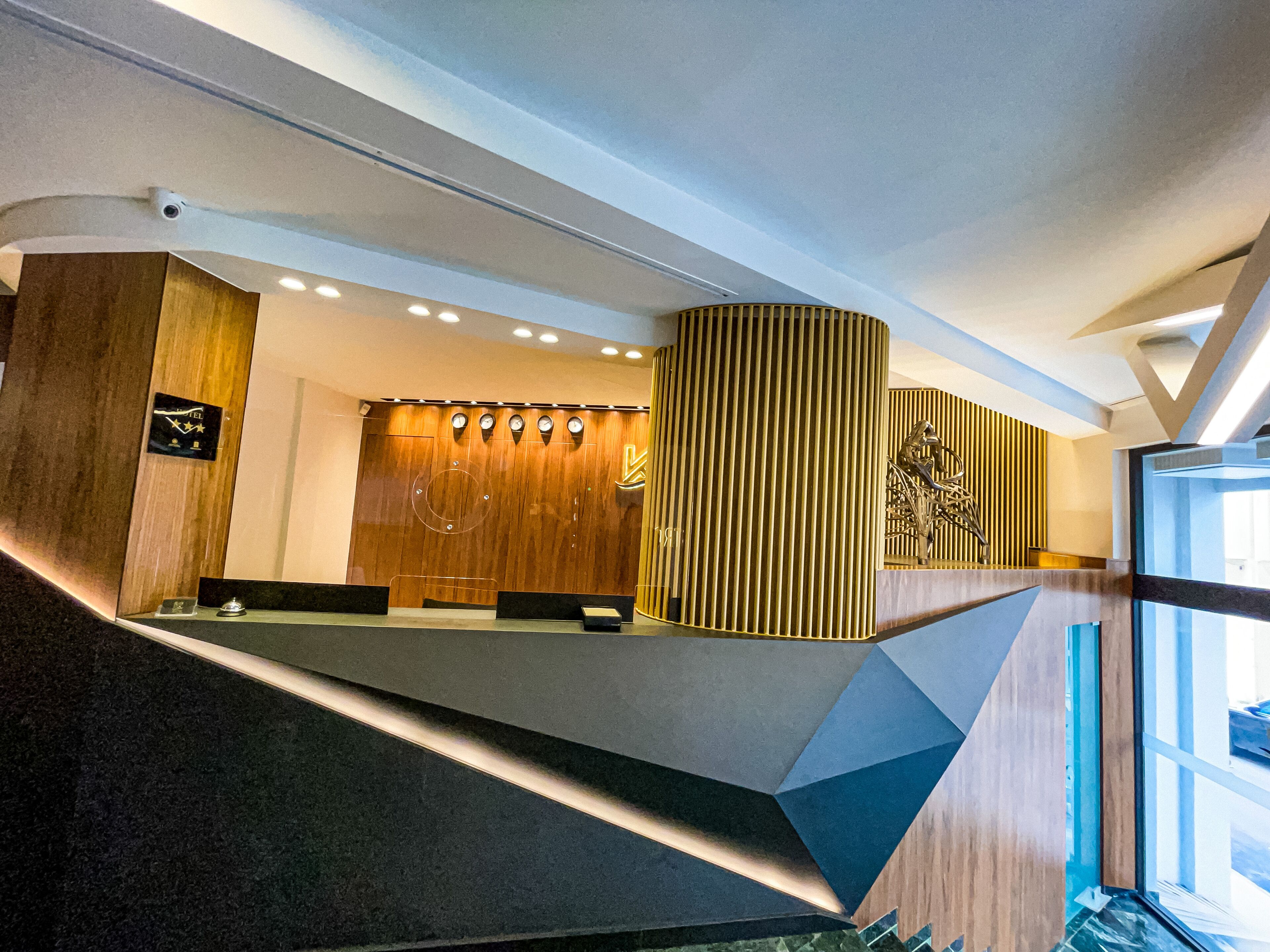 A modern reception area with a wooden reception desk, a large vertical panel with vertical lines, and a sculpture on the wall.
