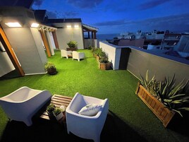 Um terraço no telhado com grama artificial, cadeiras brancas e vasos com plantas.