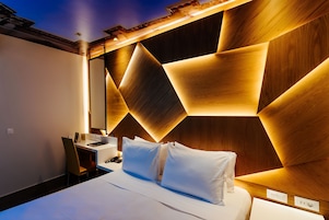 Quarto de hotel moderno com cabeceira de madeira geométrica, cama bem arrumada, escrivaninha com cadeira e pequena mesa com luminária.