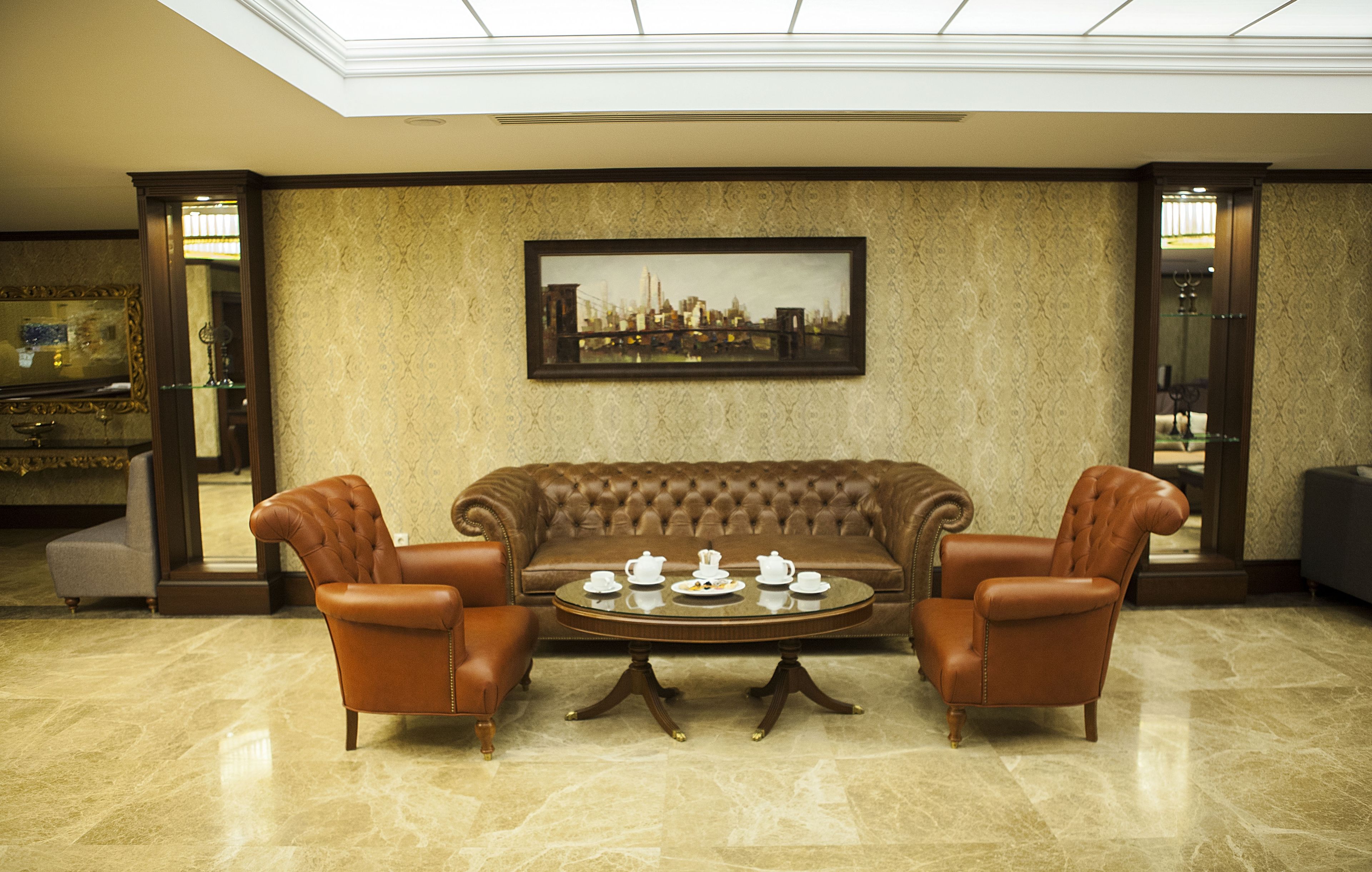 A lobby with a brown leather sofa, two leather armchairs, a round coffee table with teacups, and a framed cityscape painting on the wall.