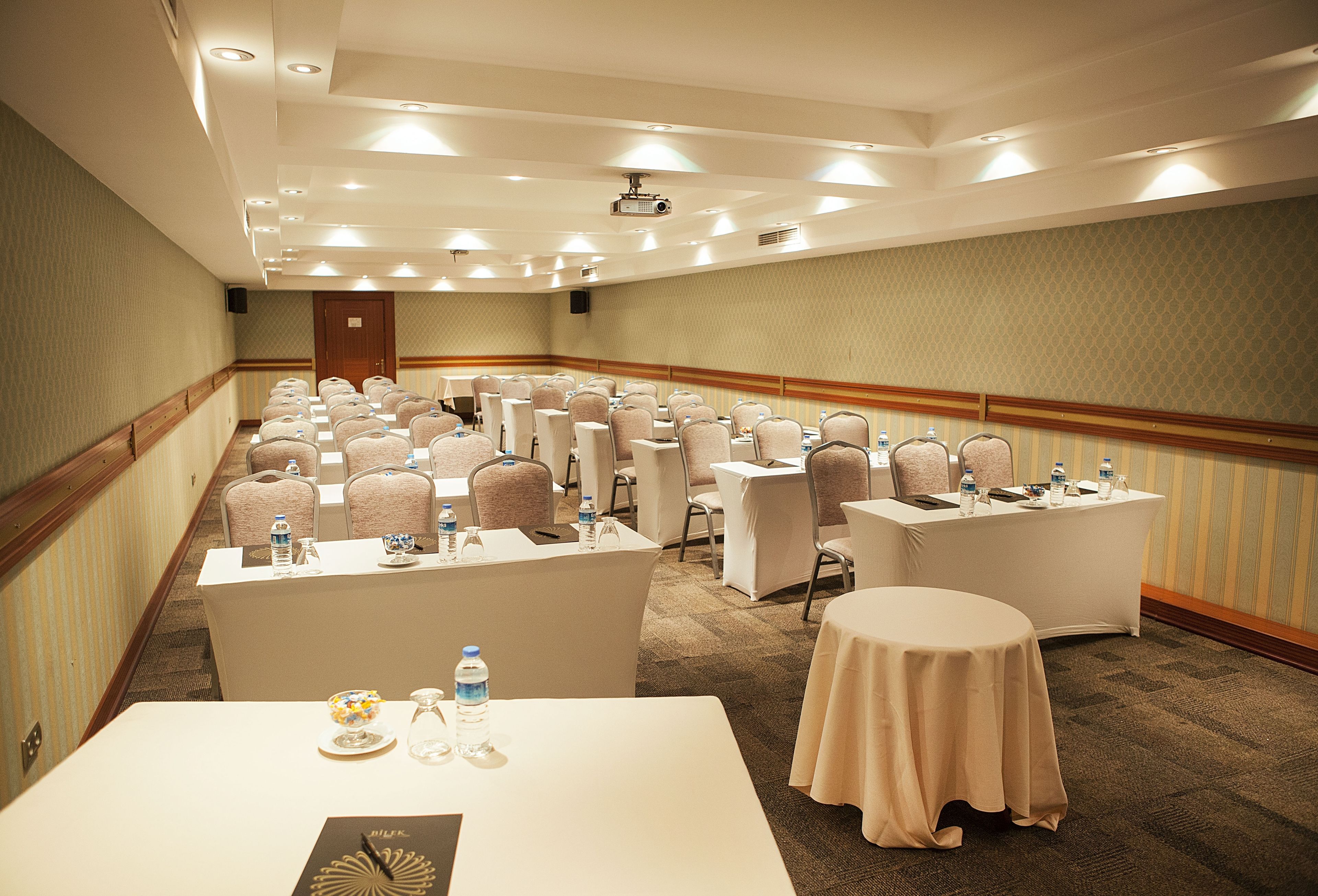 Un salón de conferencias con filas de sillas, mesas con botellas de agua y un proyector en el techo.