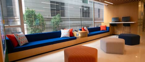 A modern office space with a long seating area, a desk with two monitors, and a large window offering a view of the outside.