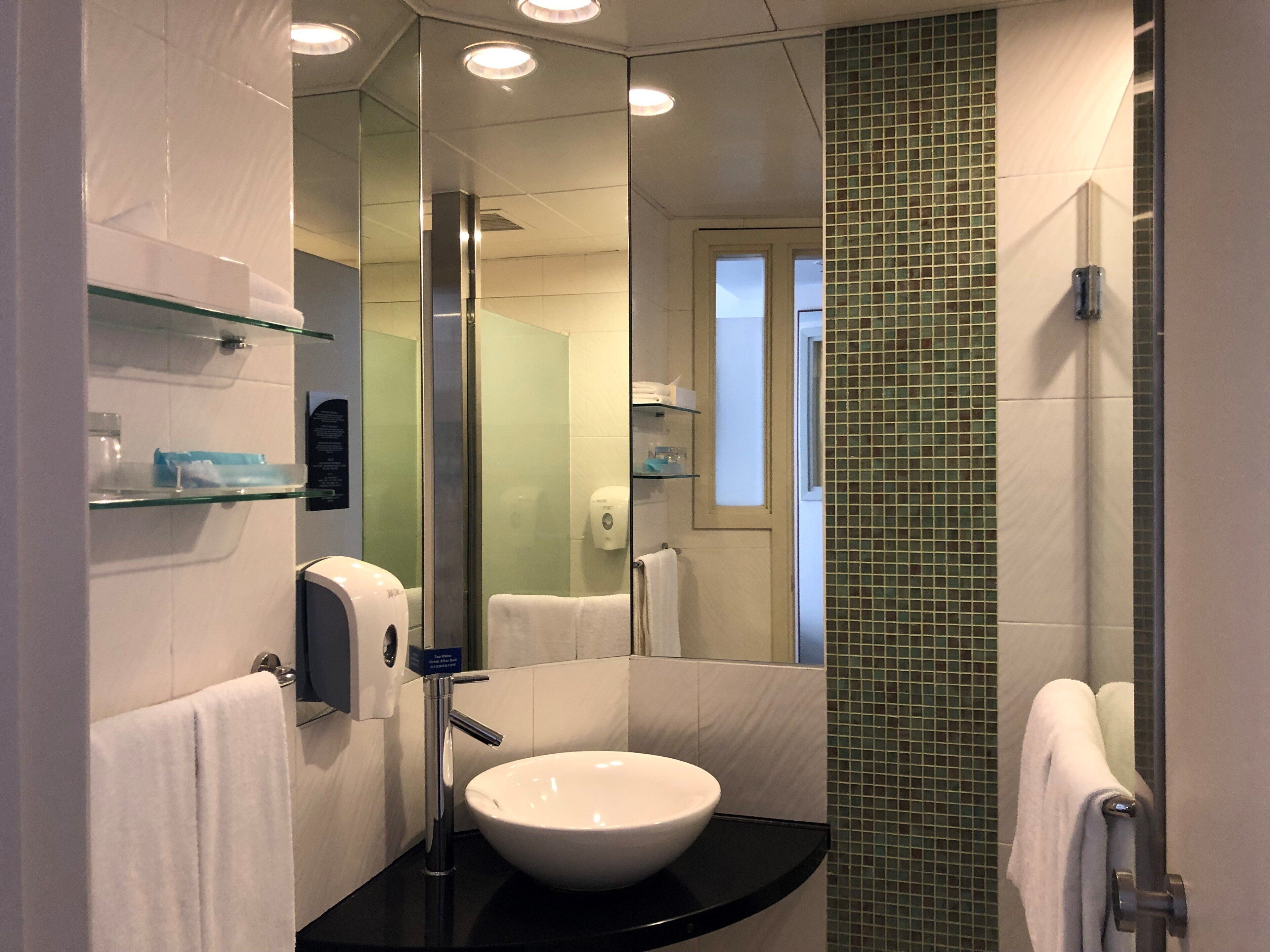 Une salle de bain moderne avec un lavabo blanc, des étagères en verre, un miroir et un mur recouvert de carreaux verts.
