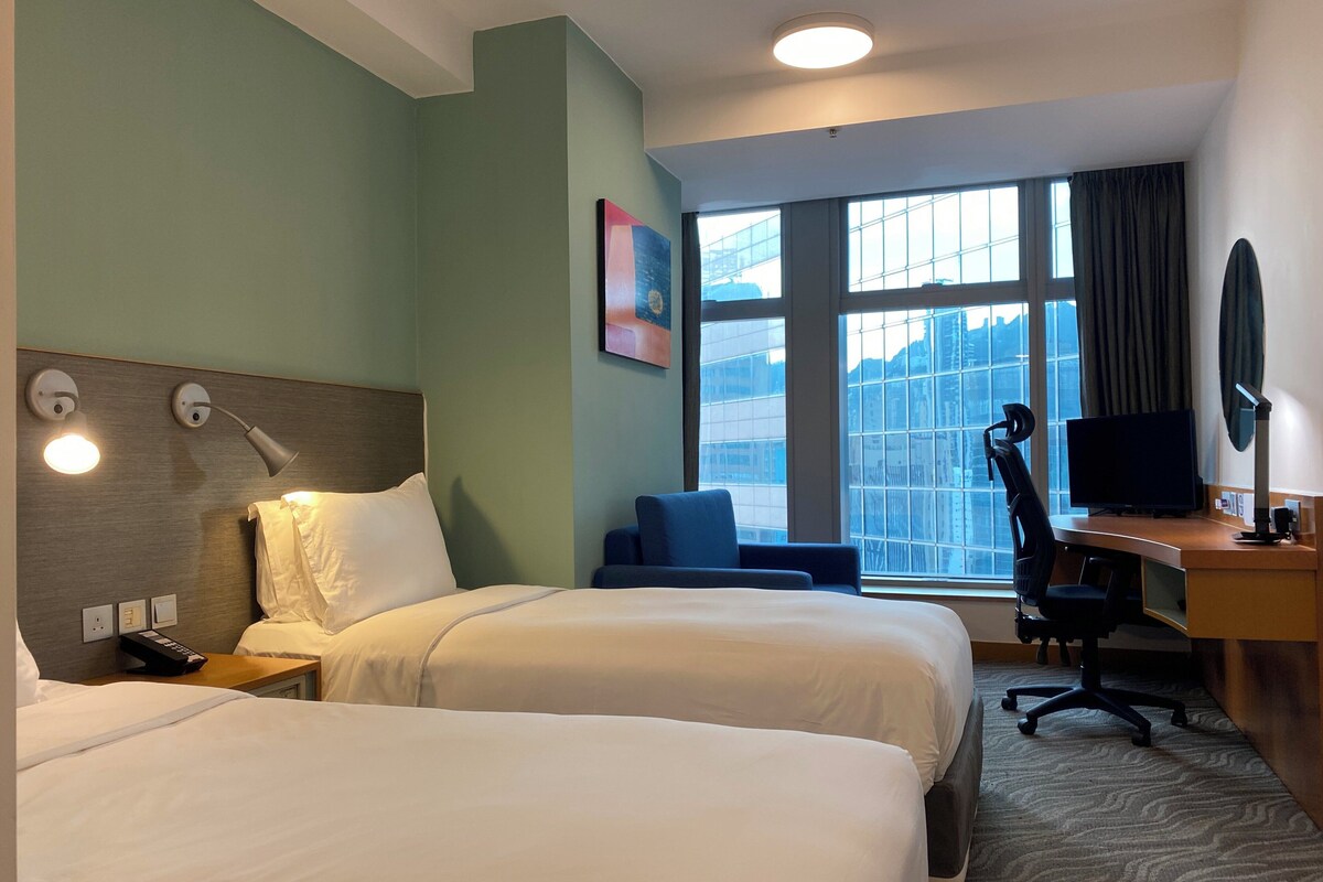 A hotel room with two beds, a desk with a computer, a chair, and a large window with a city view.