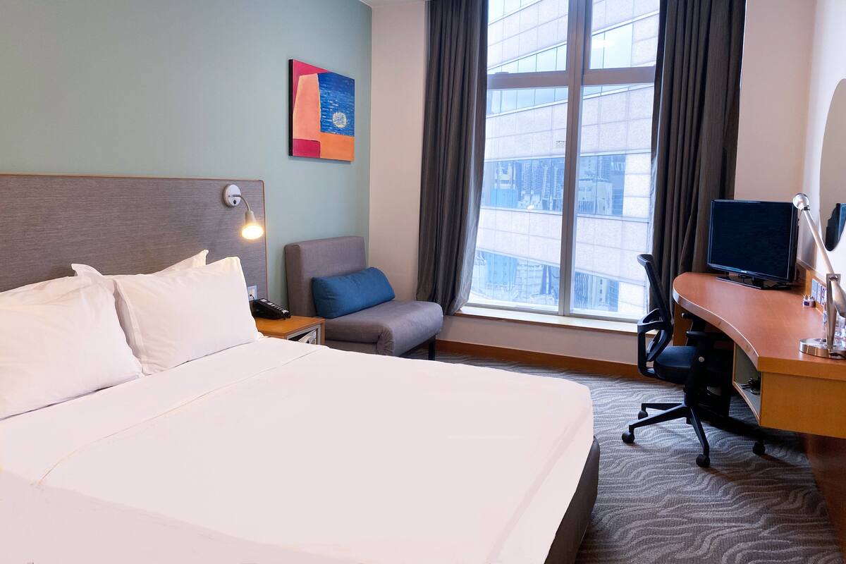 Una habitación de hotel con una cama grande, un escritorio con computadora, una silla, una lámpara y una ventana con vista a la ciudad.