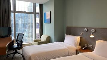 A hotel room with two beds, a desk, a chair, a TV, and a large window with a city view.