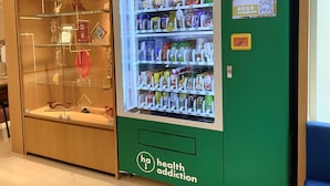 A vending machine stocked with snacks and beverages, a glass display case with trophies, and a green wall with a health addiction logo. - Holiday Inn Express Causeway Bay Hong Kong by IHG (Hong Kong)