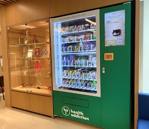 A vending machine stocked with snacks and beverages, a glass display case with trophies, and a green wall with a health addiction logo. - Holiday Inn Express Causeway Bay Hong Kong by IHG (Hong Kong)