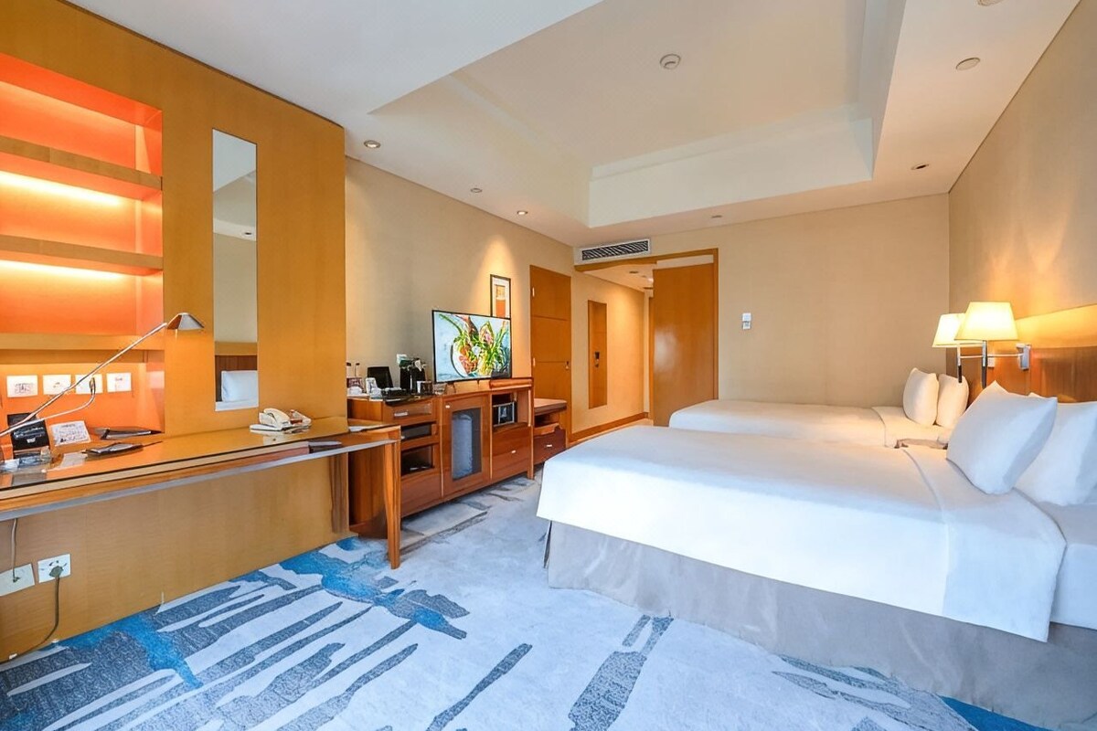 A hotel room with a desk, two beds, a large mirror, and a painting on the wall.