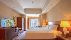 Habitación de hotel con una cama grande, un televisor sobre un mueble de madera, un escritorio con una lámpara y vistas a la ciudad a través de la ventana.