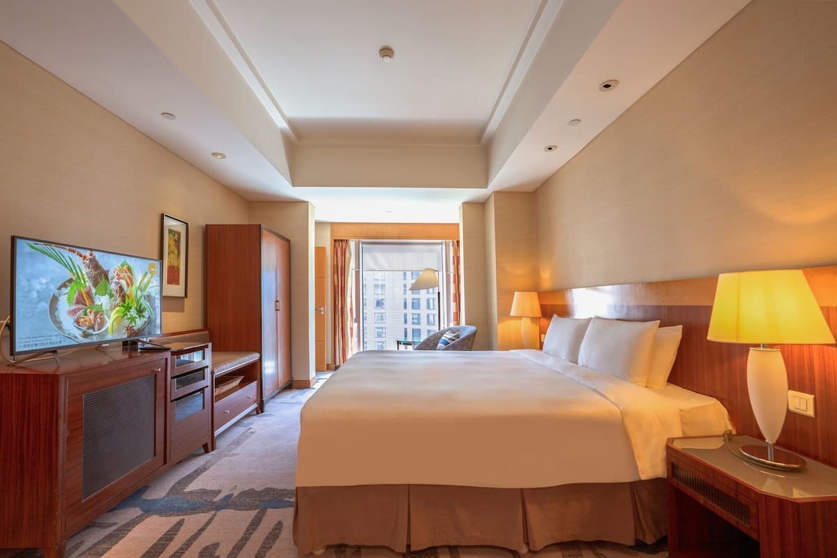 A hotel room with a large bed, a television on a wooden cabinet, a desk with a lamp, and a view of the city through the window.