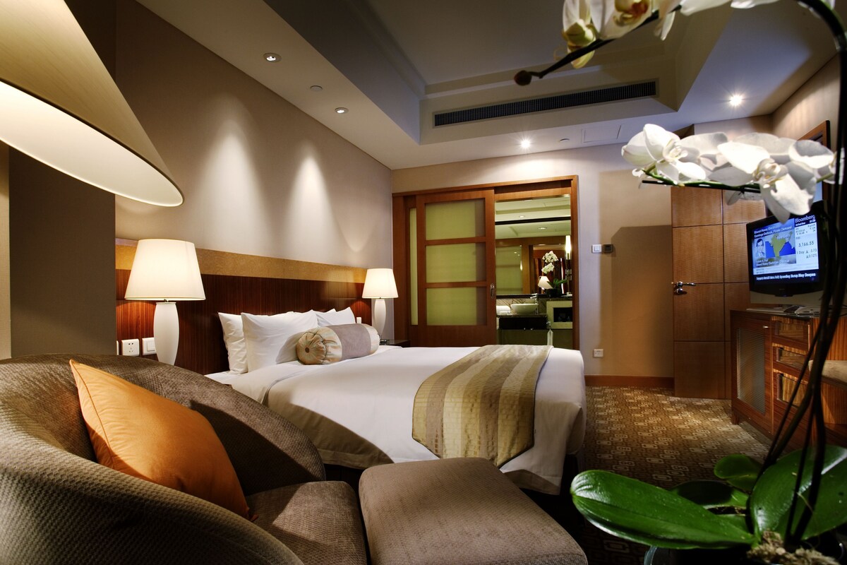 Habitación de hotel con cama, dos lámparas de noche, televisor, planta y vistas a un área de cocina.