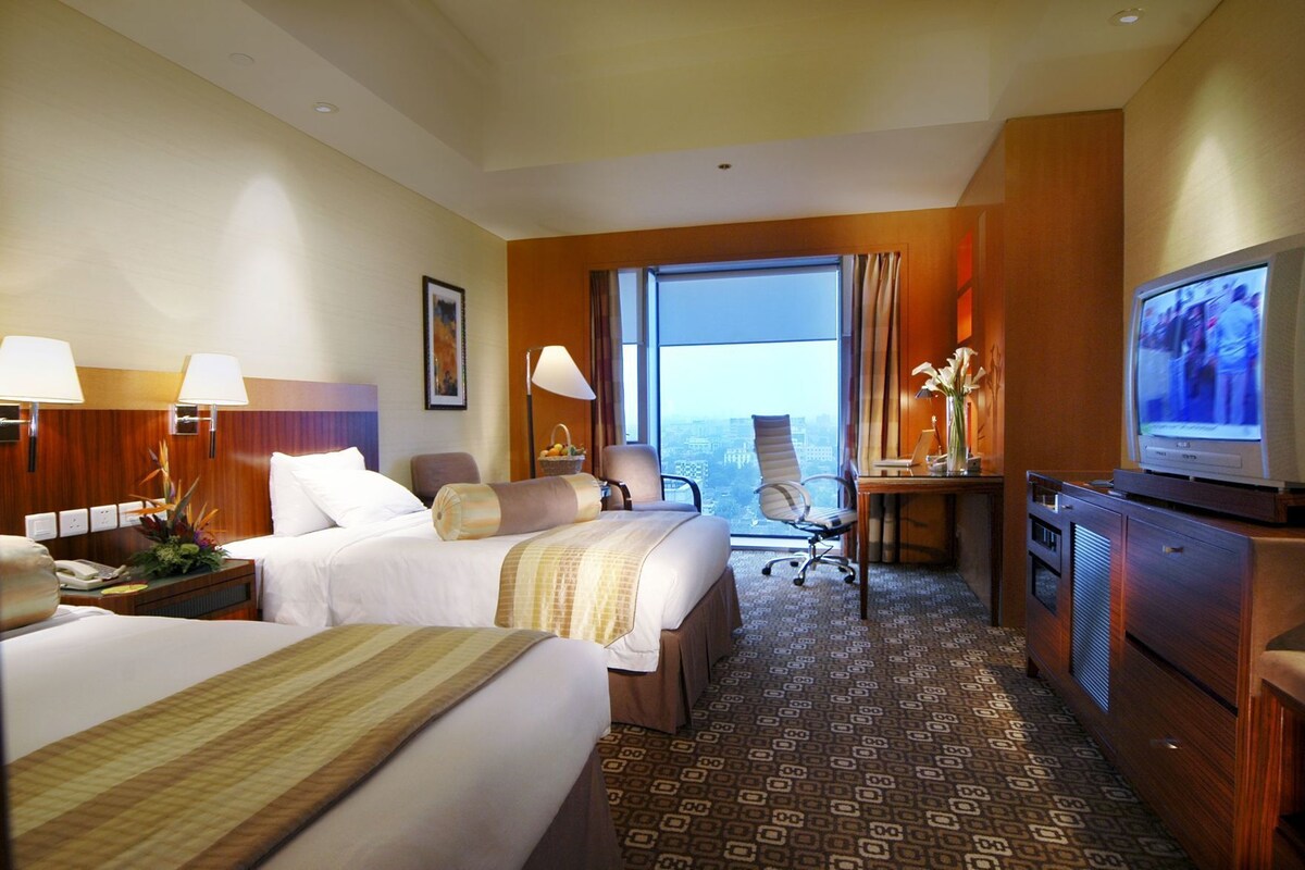 Habitación de hotel con dos camas, un escritorio, televisor y ventana grande con vistas a la ciudad.