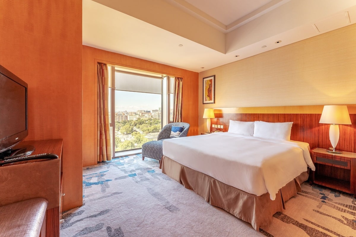 A hotel room with a large bed, a television, a chair, a desk, and a window with a city view.