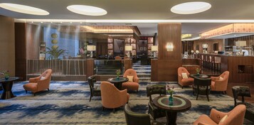 Vestíbulo de hotel con recepción, zona de estar con sillas y mesas, y un amplio ventanal con detalles decorativos.