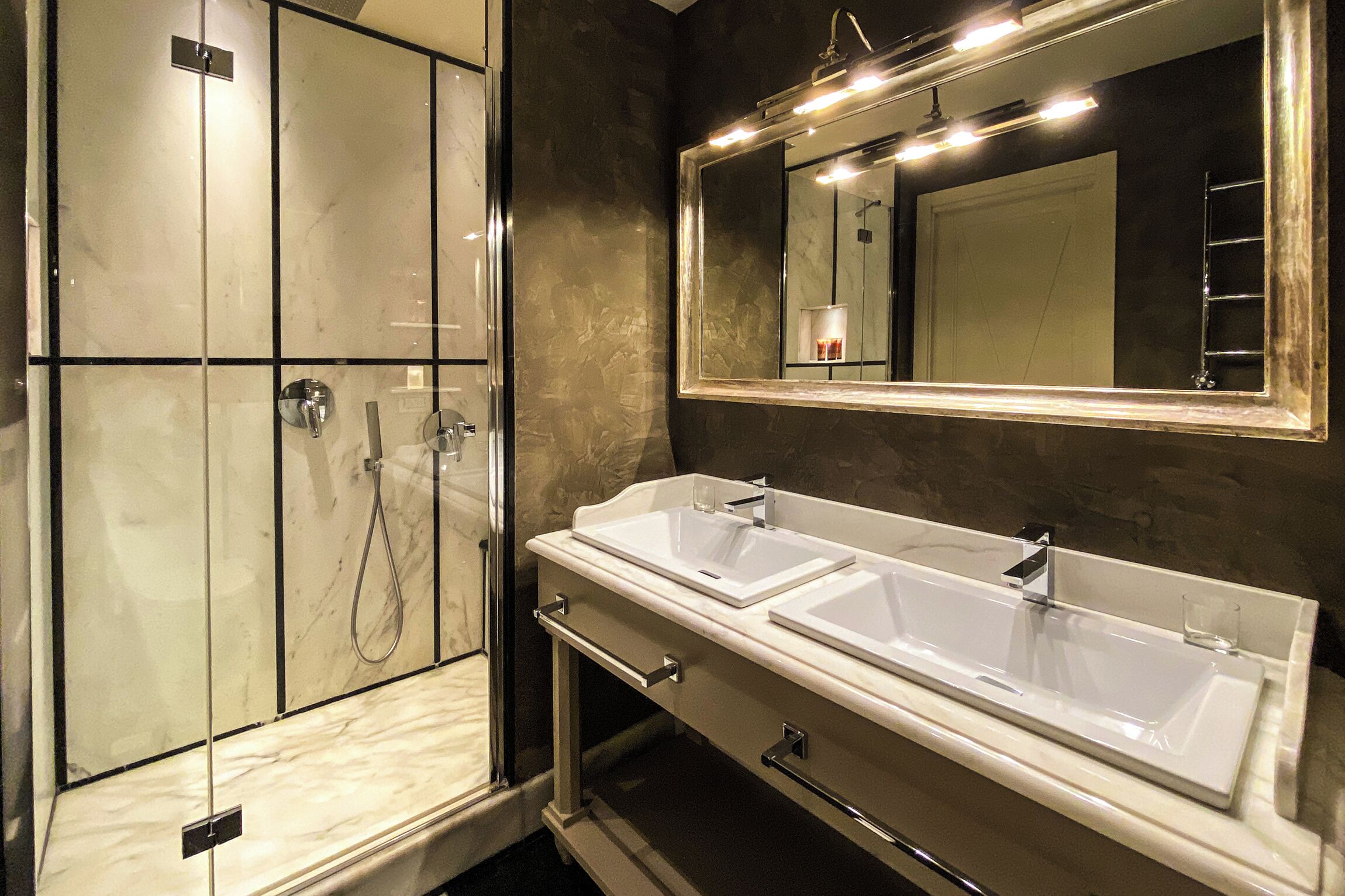 Un bagno con un grande specchio, due lavandini, una doccia e un portasciugamani.