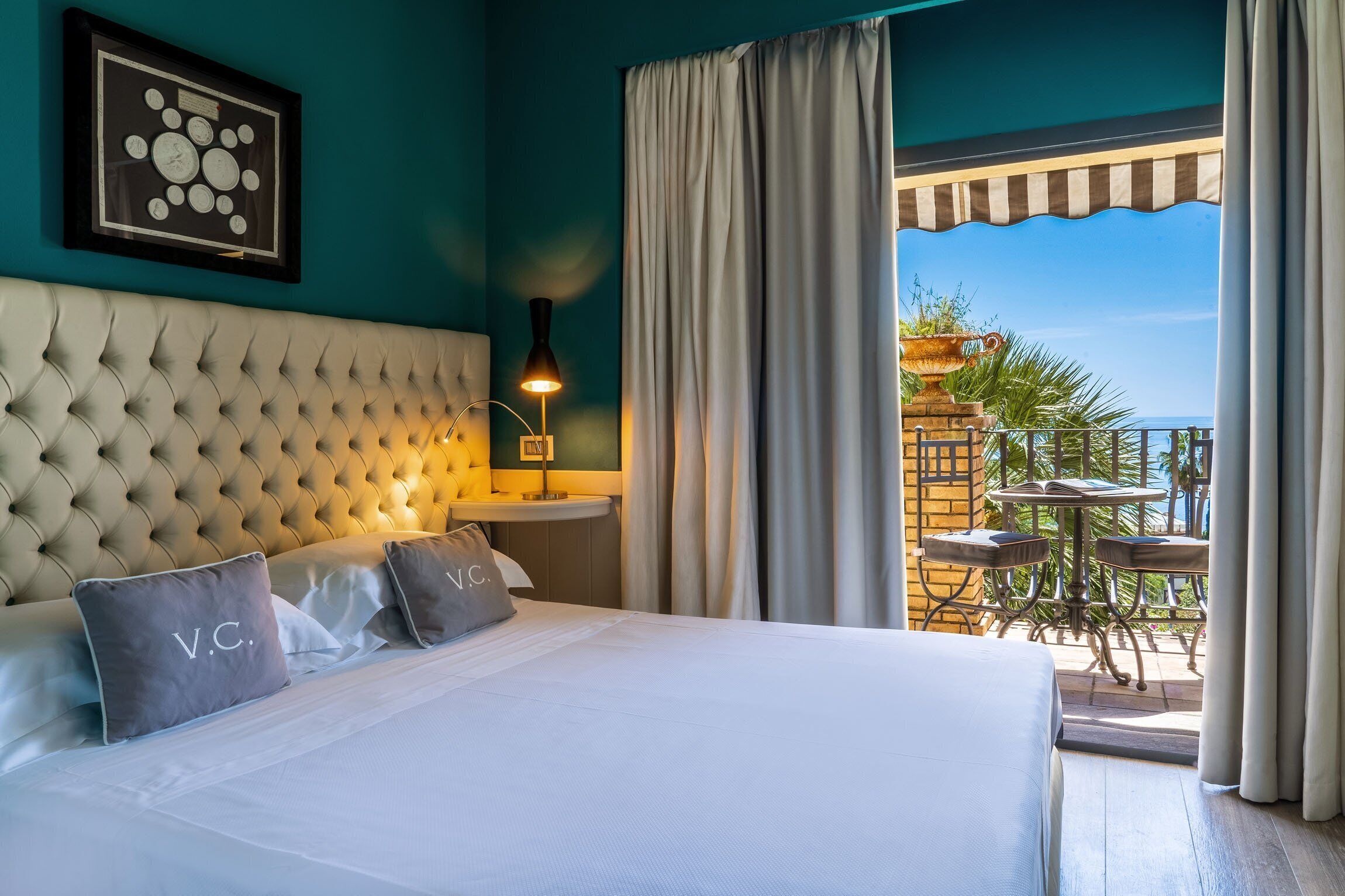 A hotel room with a tufted headboard, a framed clock on the wall, a bedside table with a lamp, and a balcony with a table and chairs.