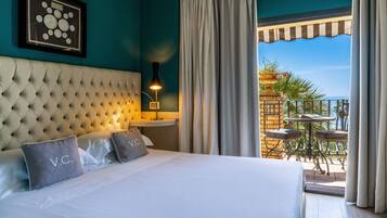 A hotel room with a tufted headboard, a framed clock on the wall, a bedside table with a lamp, and a balcony with a table and chairs.