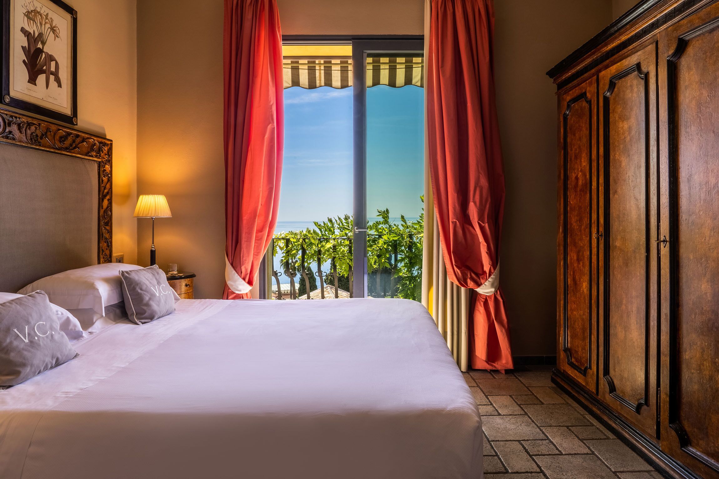 A hotel room with a bed, a nightstand, a lamp, a wardrobe, and a view of the outdoors.