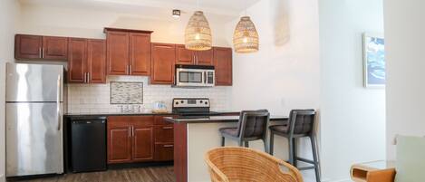 A modern kitchen with wooden cabinets, stainless steel appliances, and a central island. There are wicker chairs and a bar stool near the kitchen counter.