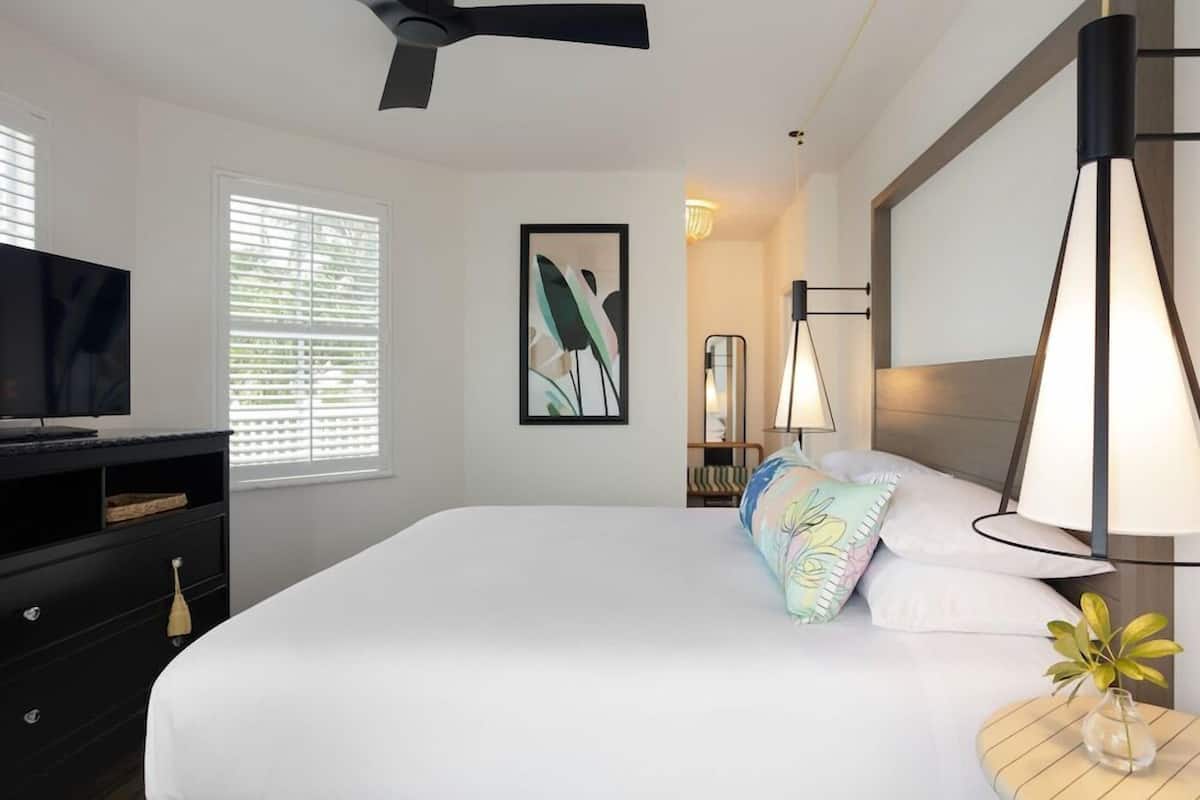 Habitación de hotel con cama, escritorio con televisor, ventilador de techo, cuadro enmarco, lámpara y una pequeña mesa con un florero.