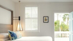 A bedroom with two beds, a window with blinds, a framed picture on the wall, and a view of the outside balcony. - Fitch Lodge - Key West Historic Inns (Key West)
