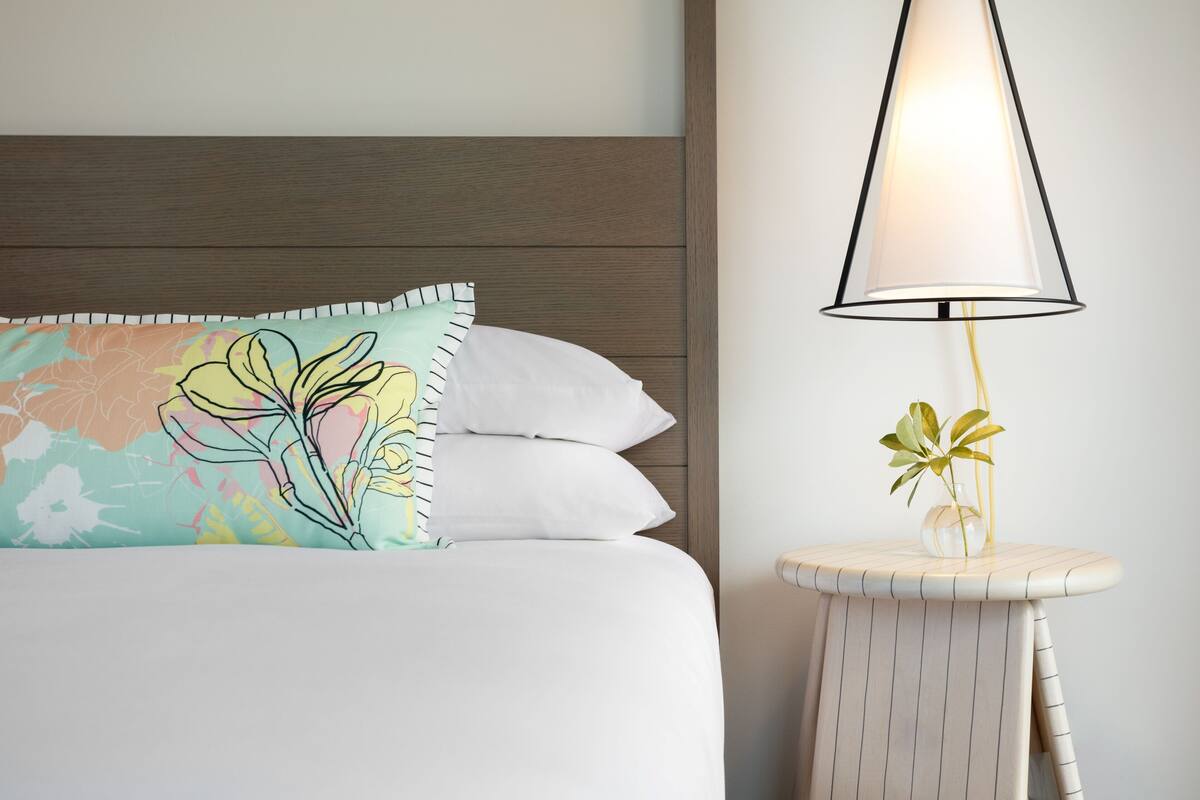 Una cama bien hecha con ropa blanca y una almohada colorida, una lámpara moderna sobre una mesita de noche y un cabecero de madera.