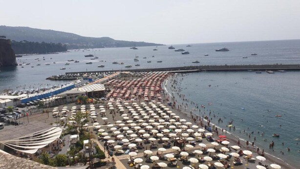 A beach with numerous umbrellas and people, a pier with boats, and a mountainous backdrop.