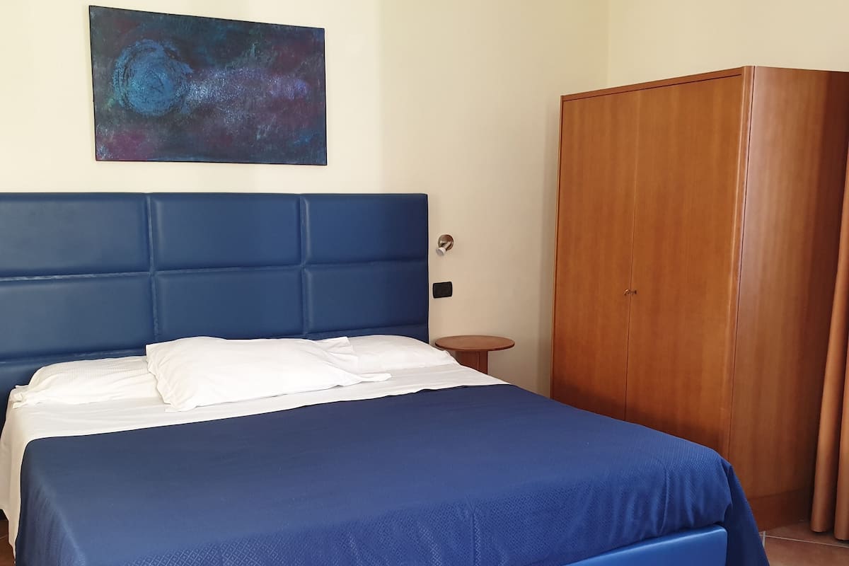 A bedroom with a blue bed, white pillows, a wooden wardrobe, and a small round table.