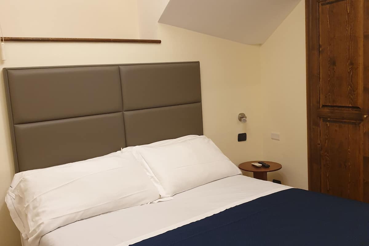 A bedroom with a bed, a wooden wardrobe, a small bedside table, and a wall-mounted light switch.