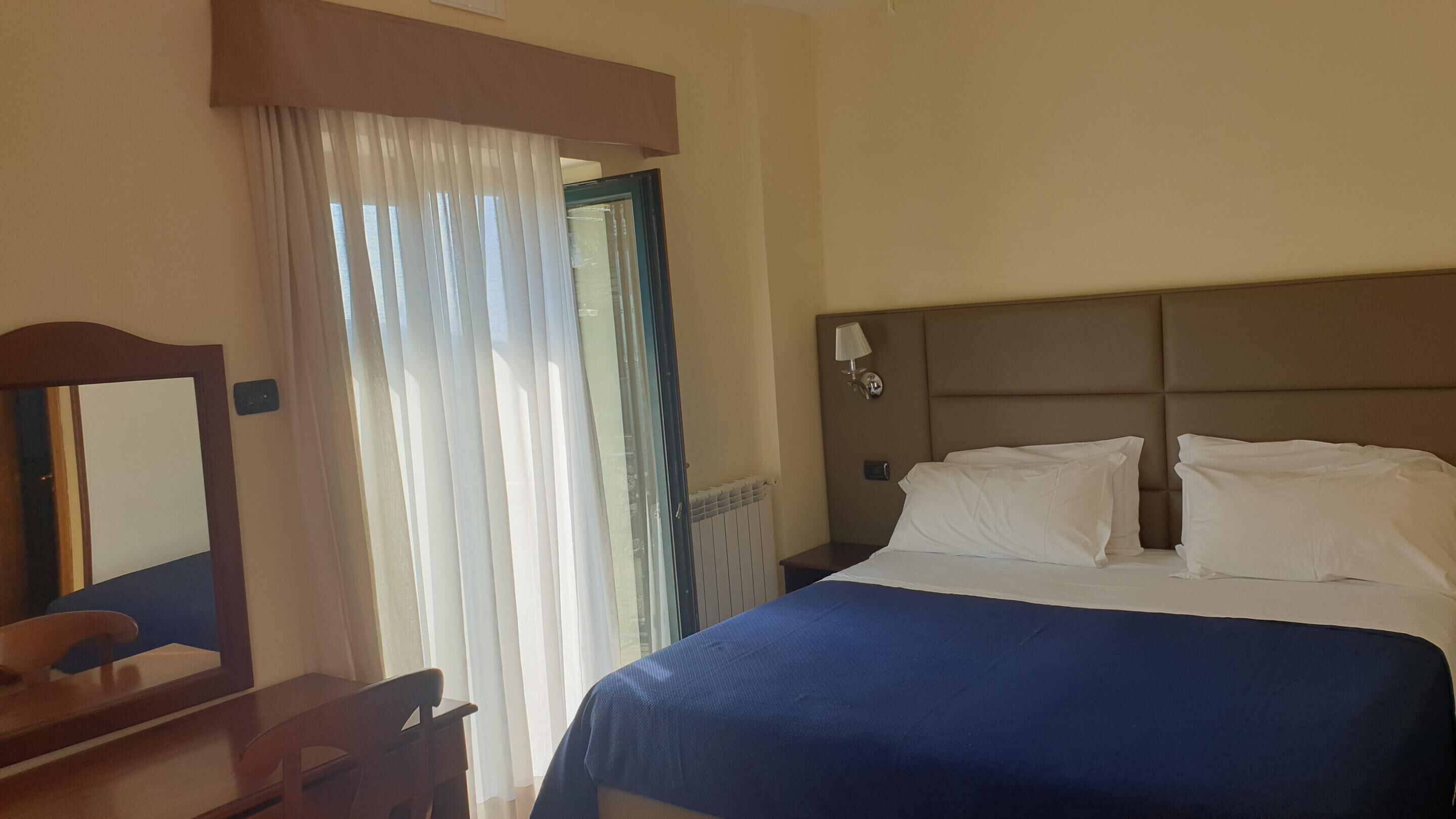 Una habitación de hotel con cama, mesita de noche, lámpara, espejo y ventana con cortinas.