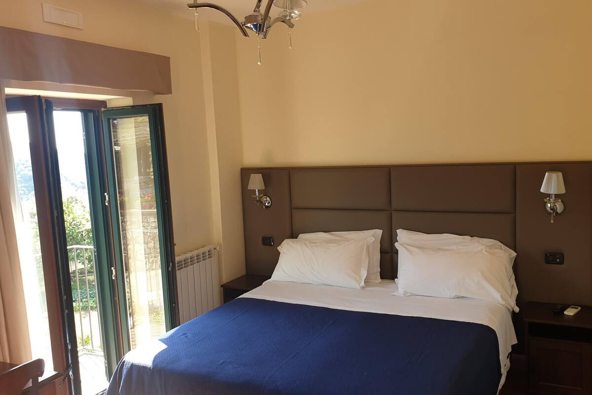 A hotel room with a bed, a nightstand, a lamp, a window with curtains, and a view of greenery outside.