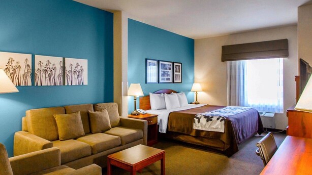 A hotel room with a blue accent wall, a bed with a purple headboard, a sofa, a small table, and a desk with a lamp. - Sleep Inn & Suites Evansville - Casper (Evansville)