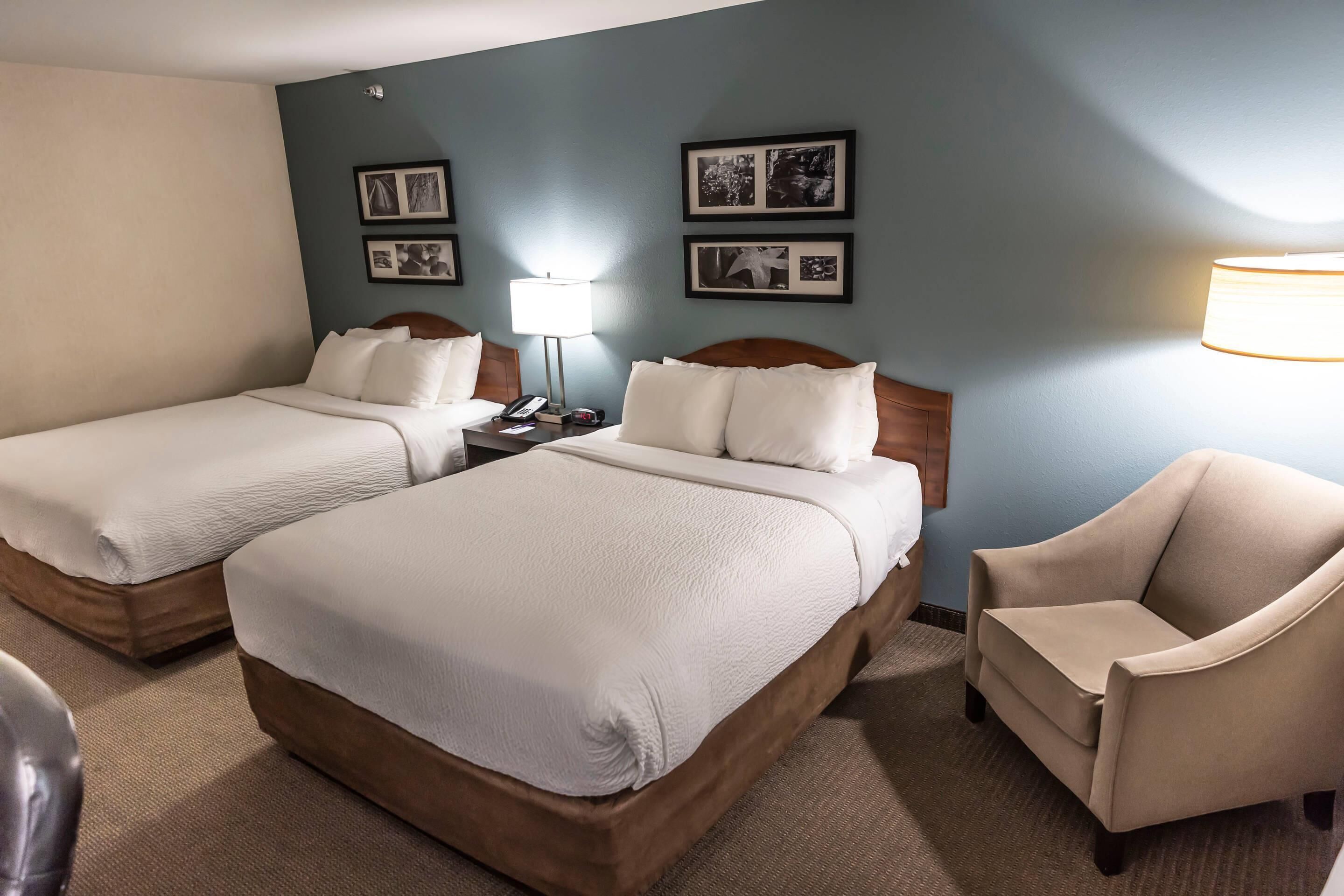 A hotel room with two beds, a chair, a nightstand with a lamp, and framed pictures on the wall.