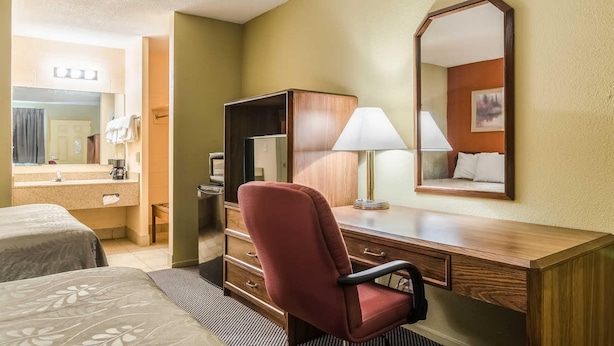 A hotel room with a wooden desk, a red chair, a large mirror, and a bed with a patterned bedspread. - Executive Inn (Dodge City)