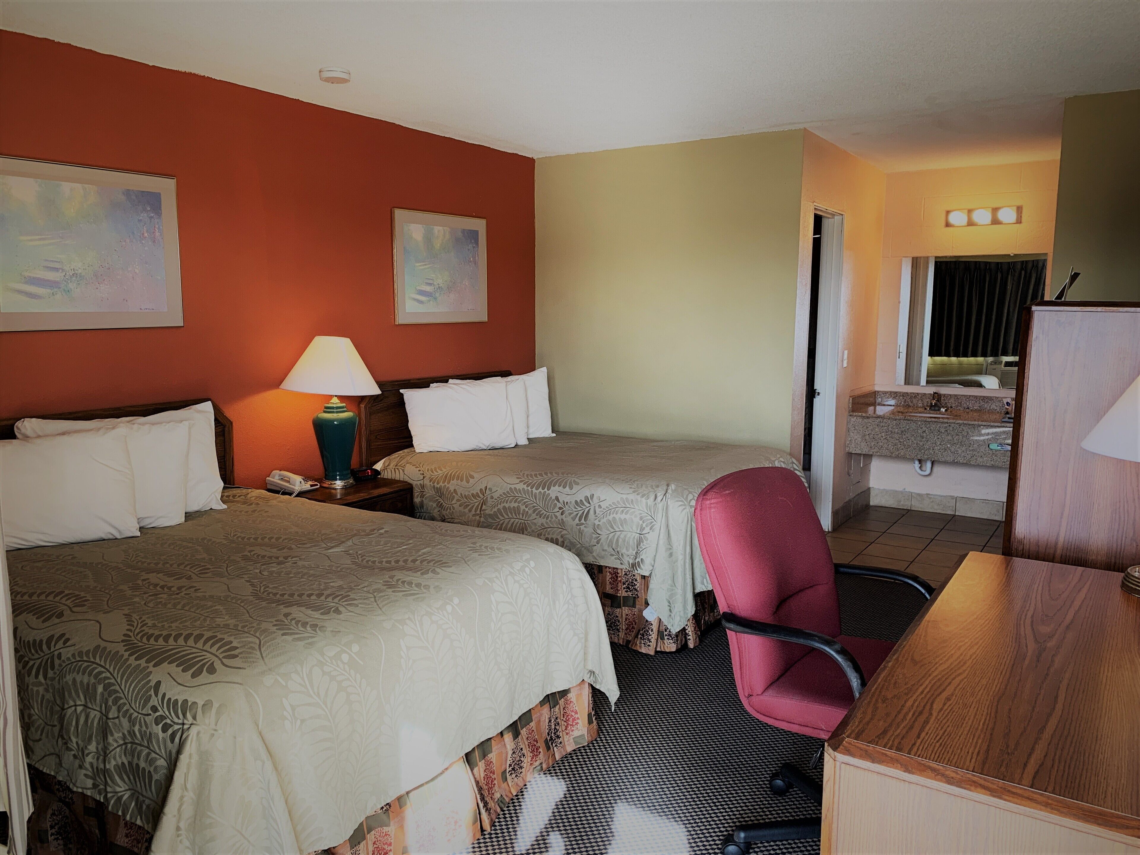 A hotel room with two beds, a desk, a chair, a lamp, and a bathroom visible through an open door.