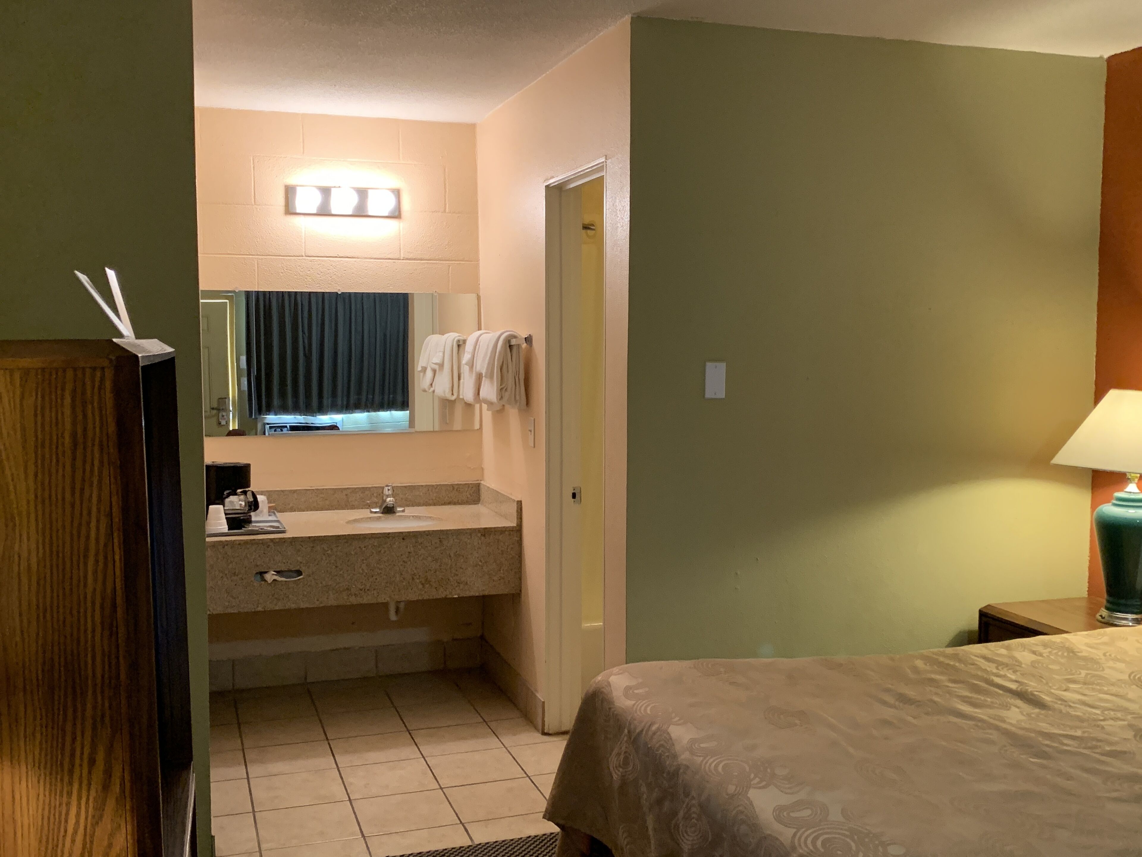 Habitació d'hotel amb llit, bany amb rentapla i mirall, i una petita taula de nit amb làmpada.