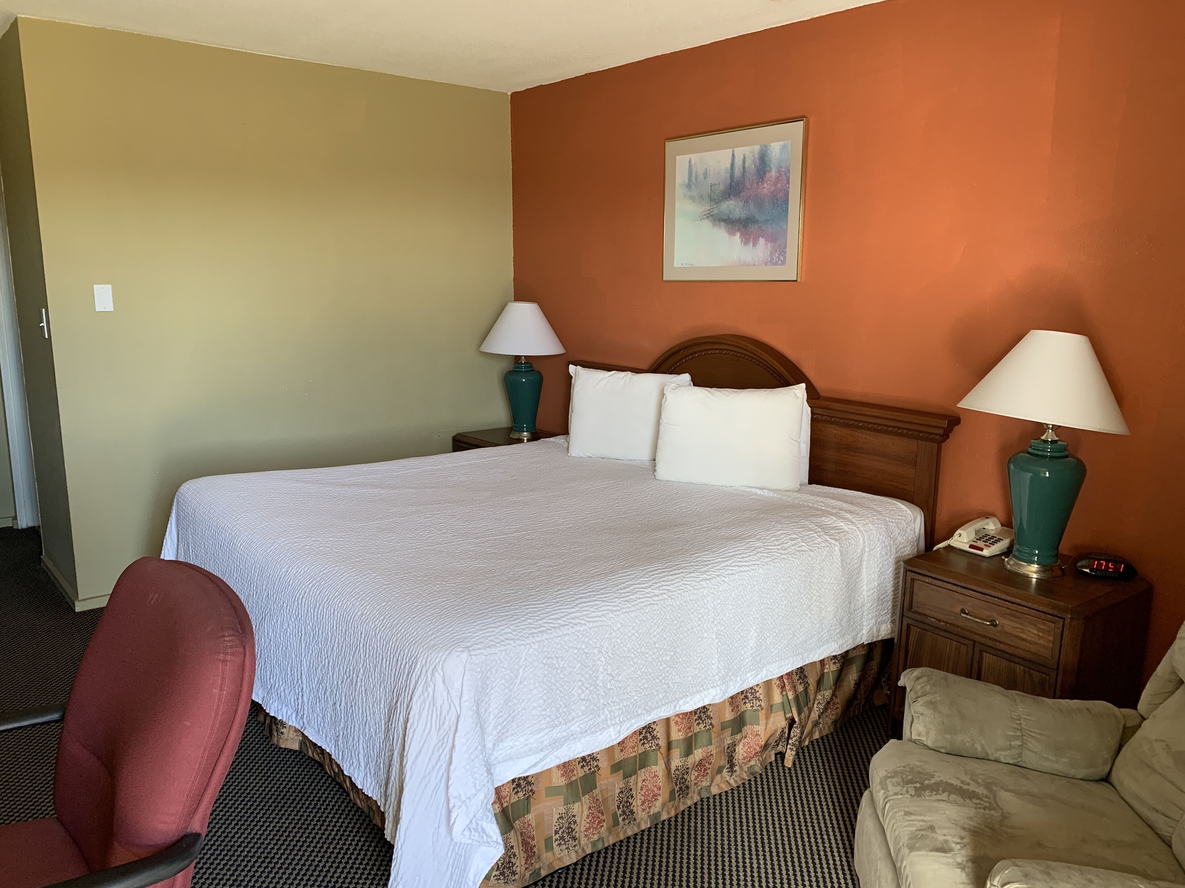 Un cuarto de hotel con una cama grande, dos mesitas de noche con lámparas, una silla y un cuadro en la pared.