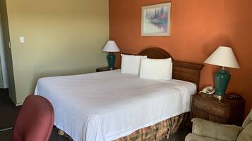 Un cuarto de hotel con una cama grande, dos mesitas de noche con lámparas, una silla y un cuadro en la pared.