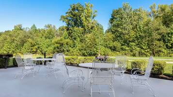 An outdoor patio with white metal chairs and a table, surrounded by lush greenery and trees.