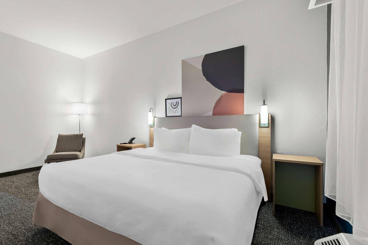 A hotel room with a large bed, a chair, a nightstand, a lamp, and a wall-mounted artwork.