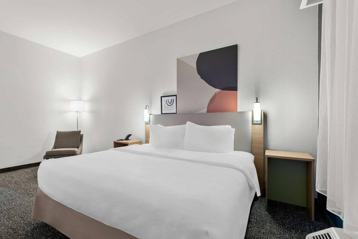 A hotel room with a large bed, a chair, a nightstand, a lamp, and a wall-mounted artwork.