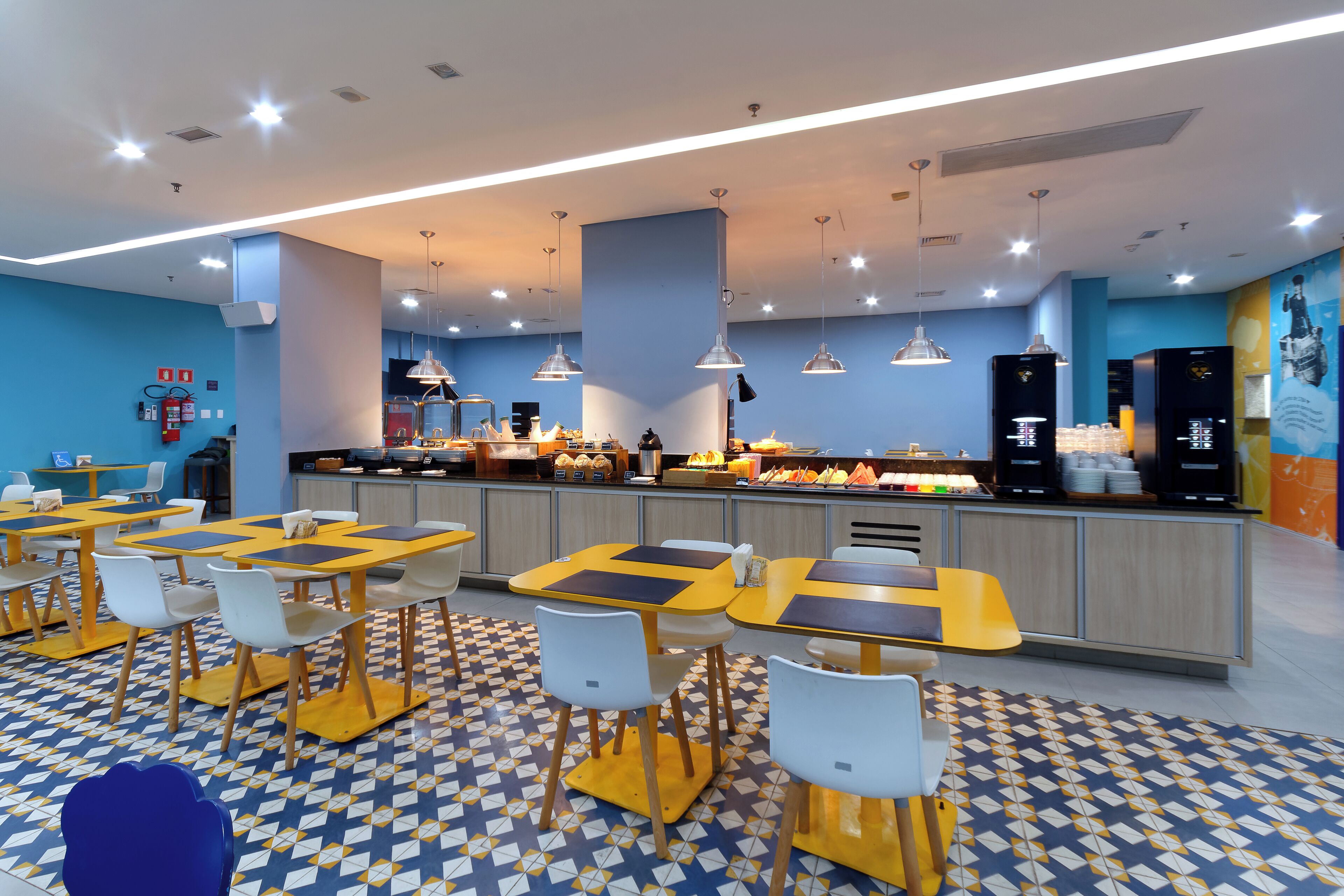 Una cafetería moderna con varias mesas y sillas, un mostrador con alimentos y iluminación colgante.