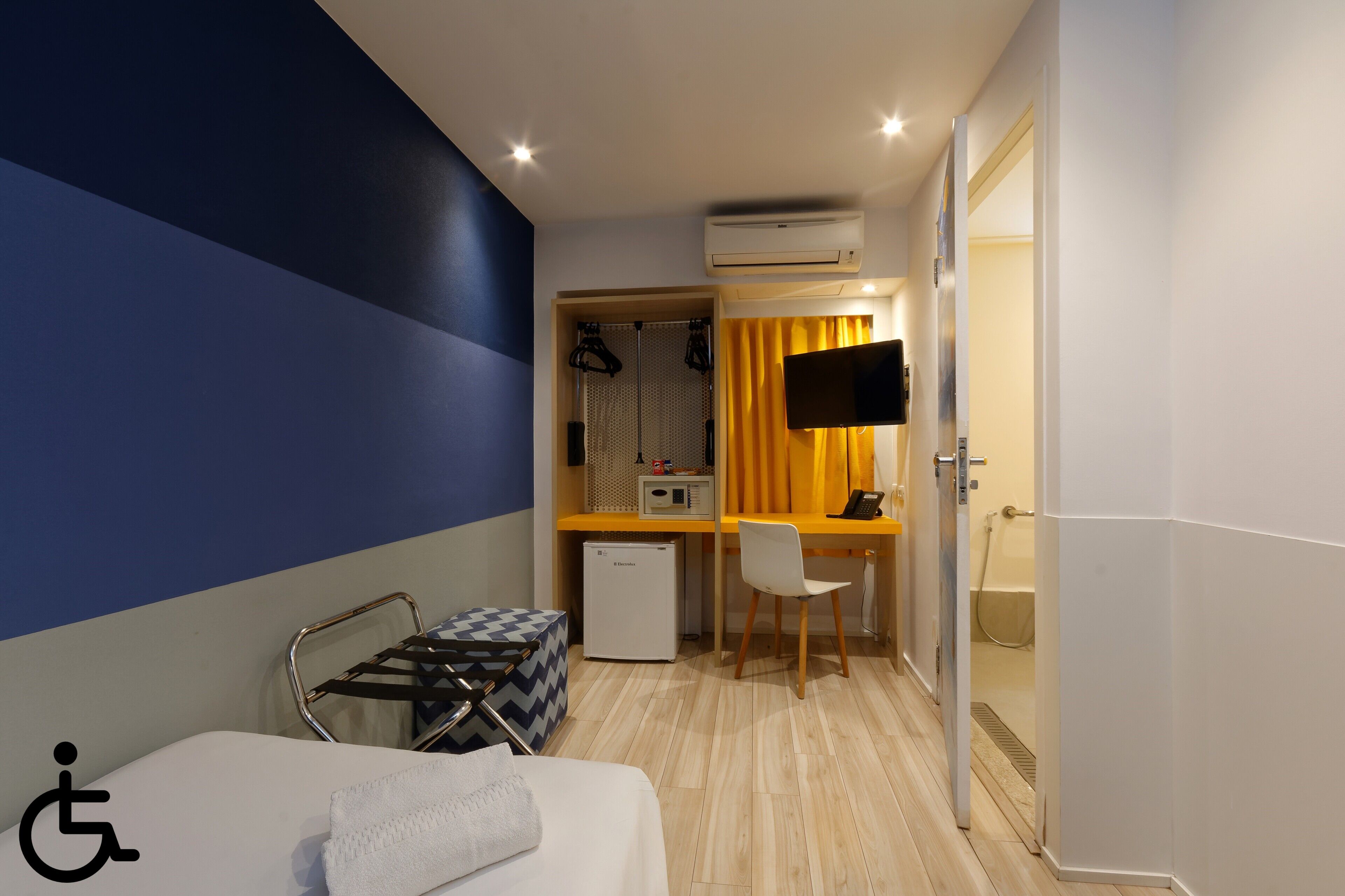 Quarto de hotel com cama, escrivaninha com cadeira, televisão, ar condicionado, mini-geladeira e chuveiro.