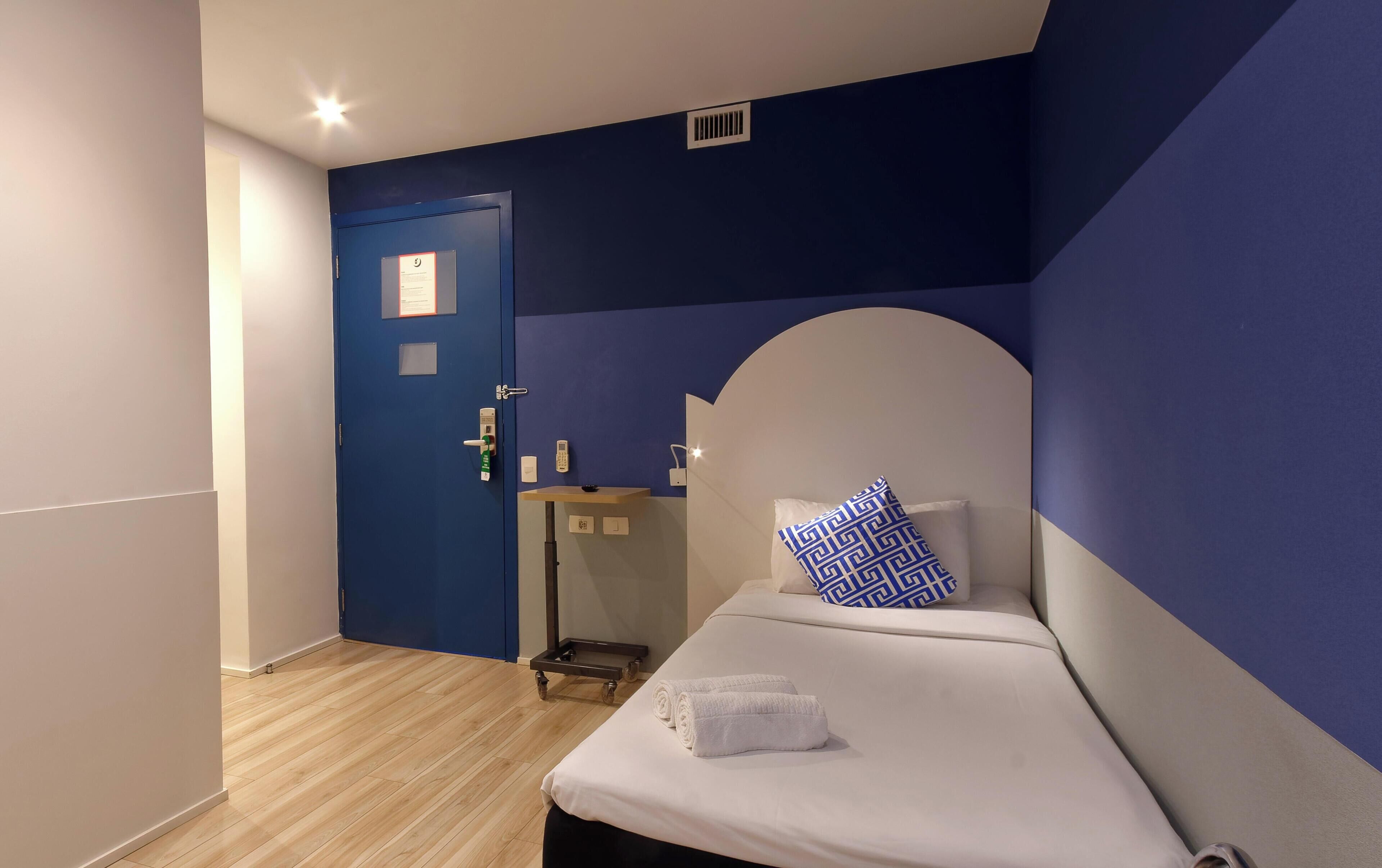 Quarto de hotel com uma parede azul decorativa, uma cama com roupa de cama branca e almofada estampada, uma mesa de cabeceira com telefone e uma porta com fechadura por cartão.